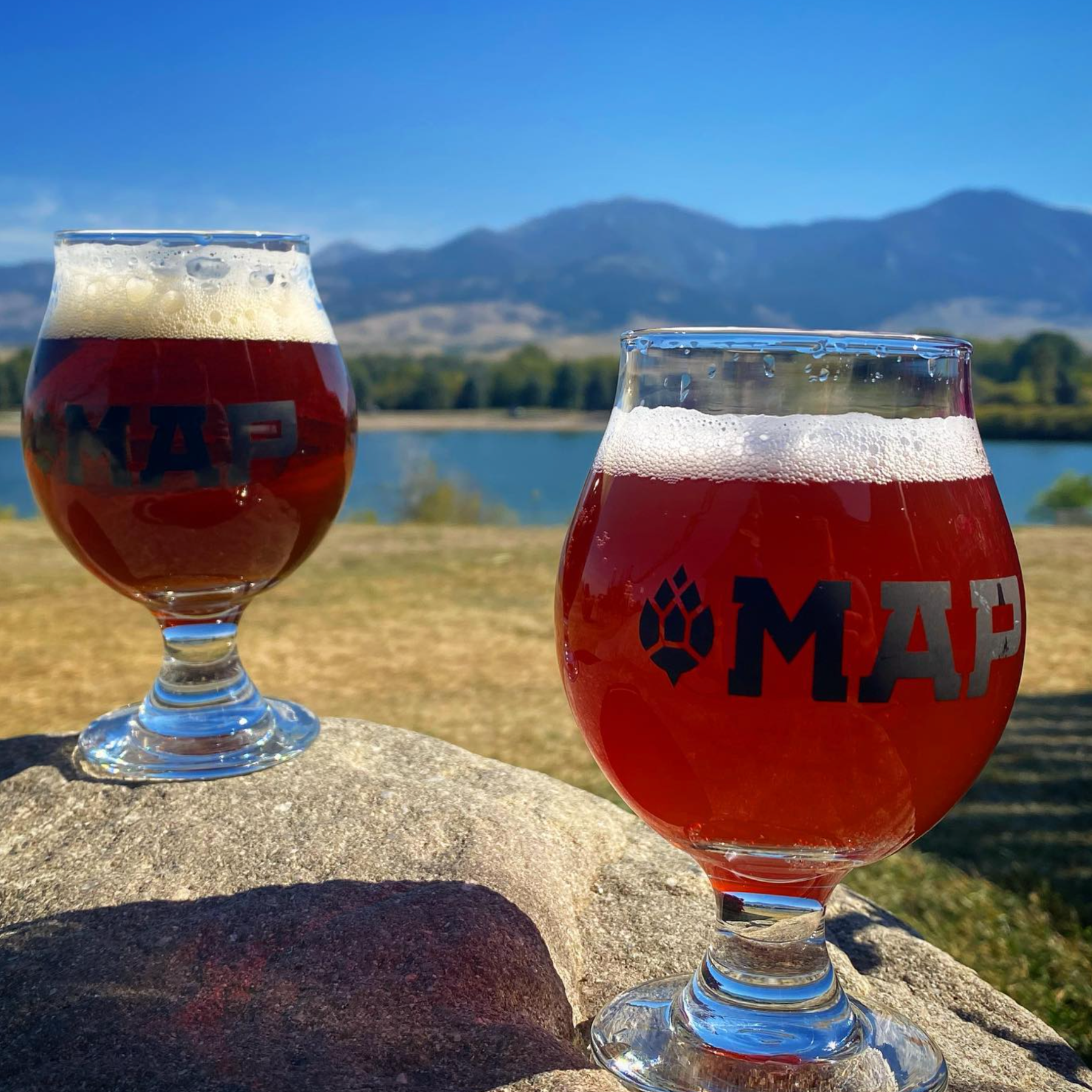 Two glasses of beer on rocks, mountains and lake in background under blue sky.
