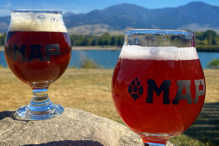 Two glasses of beer on rocks, mountains and lake in background under blue sky.