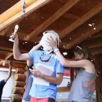 Person with paper hit by pie, holding trophy outdoors under wood structure.