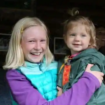 Smiling girl holding a toddler in colorful jackets indoors.