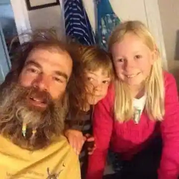Man with long beard and two smiling children indoors.