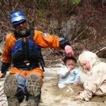 Man in orange suit with two children outdoors, seated near dry branches.