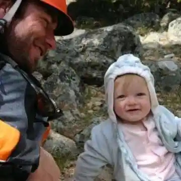 Smiling man in helmet and baby in hooded jacket outdoors.