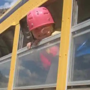 Child in pink helmet looks out school bus window smiling.